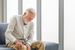 Elderly man experiencing knee pain on sofa, highlighting the need for chiropractic care for seniors at Enlightened Chiropractic.